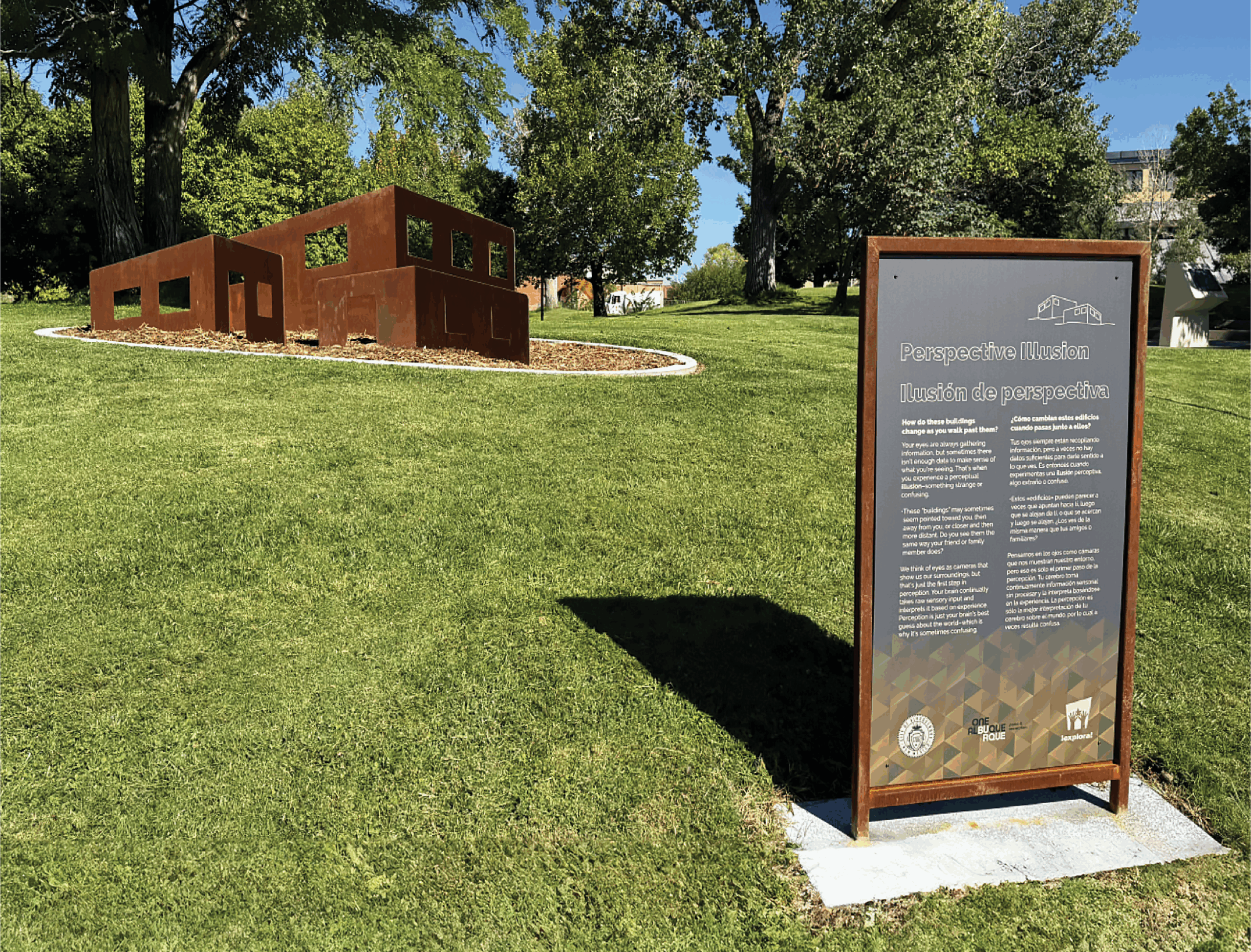 A metal sculpture made of rust-colored rectangular frames sits on a grassy hill under tall trees. In the foreground, a vertical informational sign titled ‘Perspective Illusion’ explains the installation. The sculpture is arranged so viewers can see different shapes depending on their angle